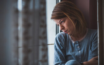 A woman wearing a blue top sits by a window with a distant, contemplative expression. Her arms are crossed over her knees, and light filters through lace curtains nearby, casting a soft glow on her face. The scene evokes a mood of introspection and melancholy.