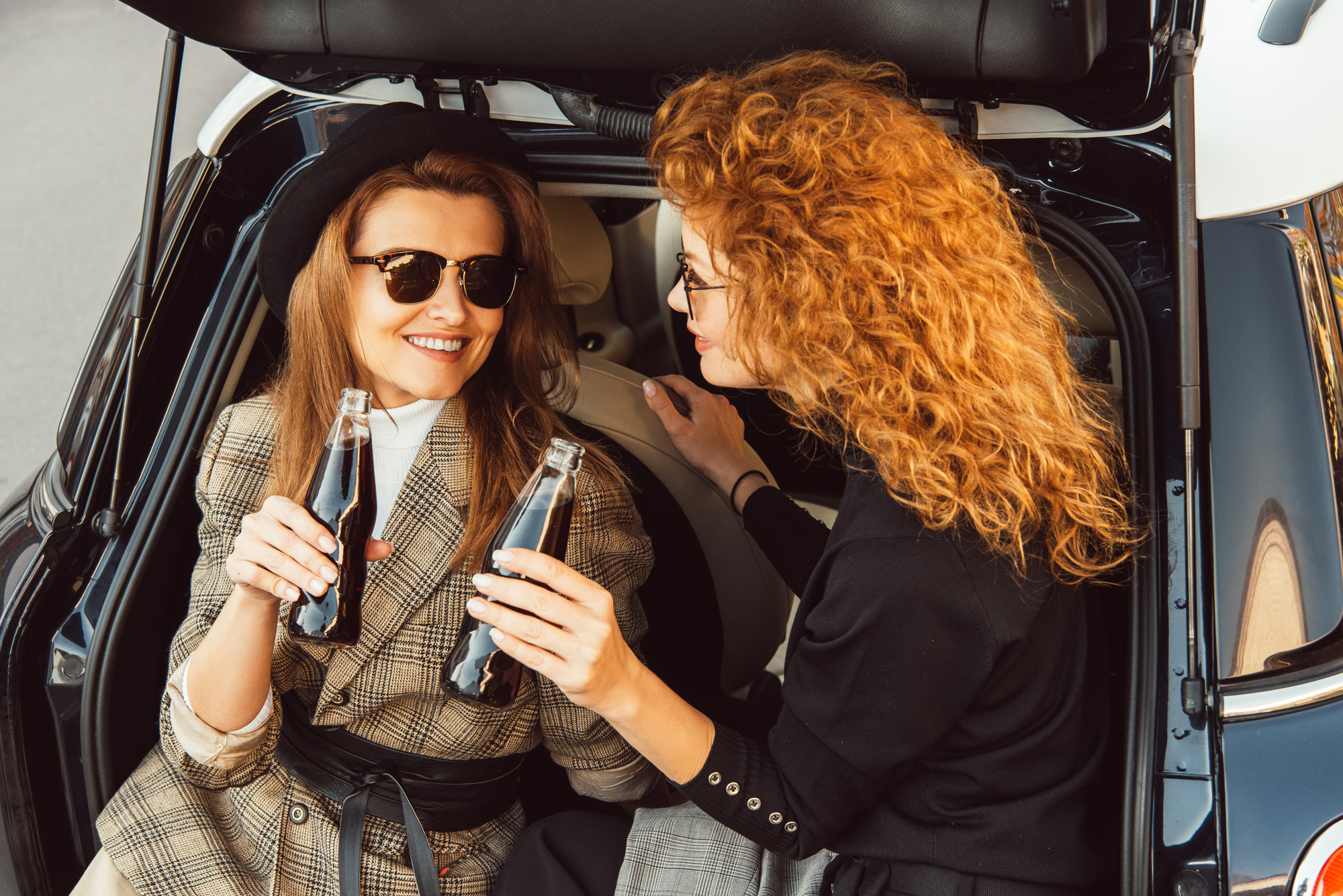 Two women sit in the open trunk of a car, smiling and holding glass bottles of soda. Both are wearing sunglasses; one has red hair and a plaid outfit, the other curly red hair and a black top. The mood is casual and cheerful.