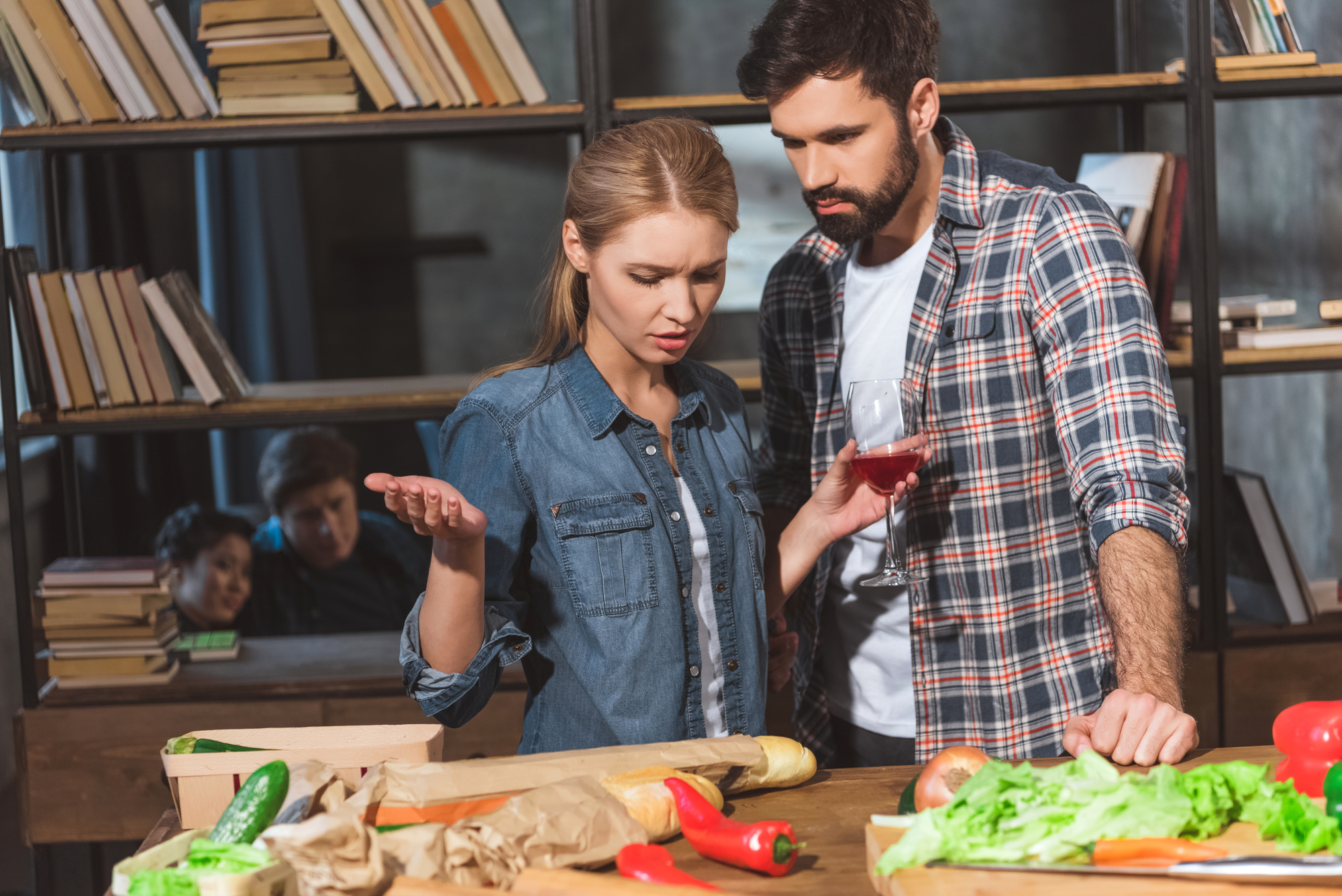 A woman gestures in confusion while a man holding a glass of red wine looks at vegetables on a table in a kitchen. They are surrounded by bookshelves filled with books, and two other people can be seen in the background, sitting and talking.