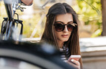 A woman with long, dark hair sits outdoors wearing large, round sunglasses. She is looking down at her smartphone with a focused expression. Part of a bicycle is visible in the foreground, and greenery can be seen in the blurred background.