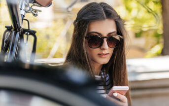 A woman with long, dark hair sits outdoors wearing large, round sunglasses. She is looking down at her smartphone with a focused expression. Part of a bicycle is visible in the foreground, and greenery can be seen in the blurred background.