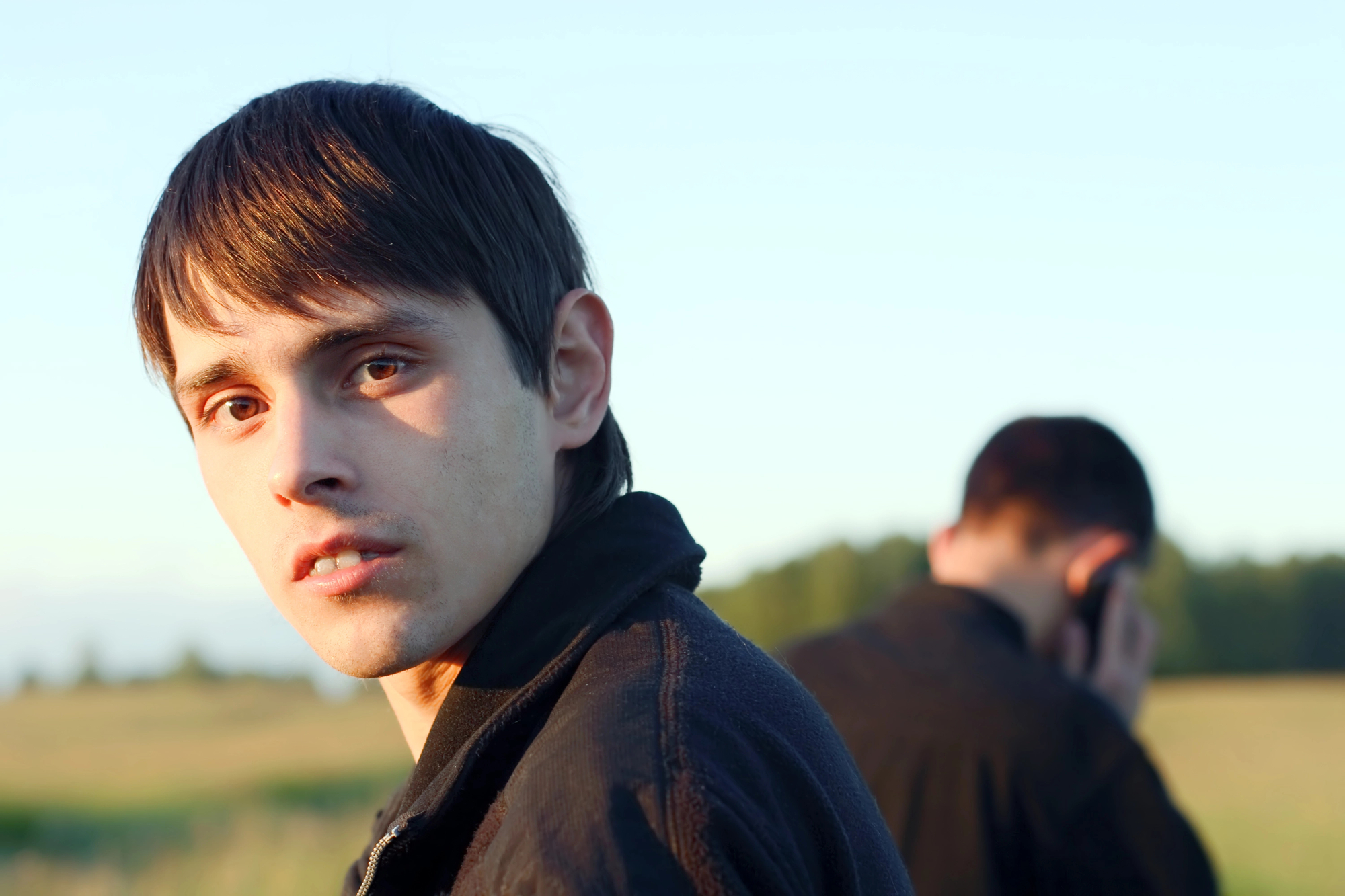 A young man with short dark hair and a serious expression looks directly at the camera in a field. Another person is in the background with their back turned, talking on a phone. Both wear dark jackets, and trees are visible in the distance.