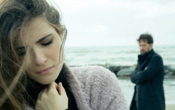 A pensive woman with long hair stands in the foreground near the shore, her eyes closed and hand near her chest. A man with a beard, wearing a dark coat, stands in the background with crossed arms, looking out at the sea. The mood is somber and reflective.