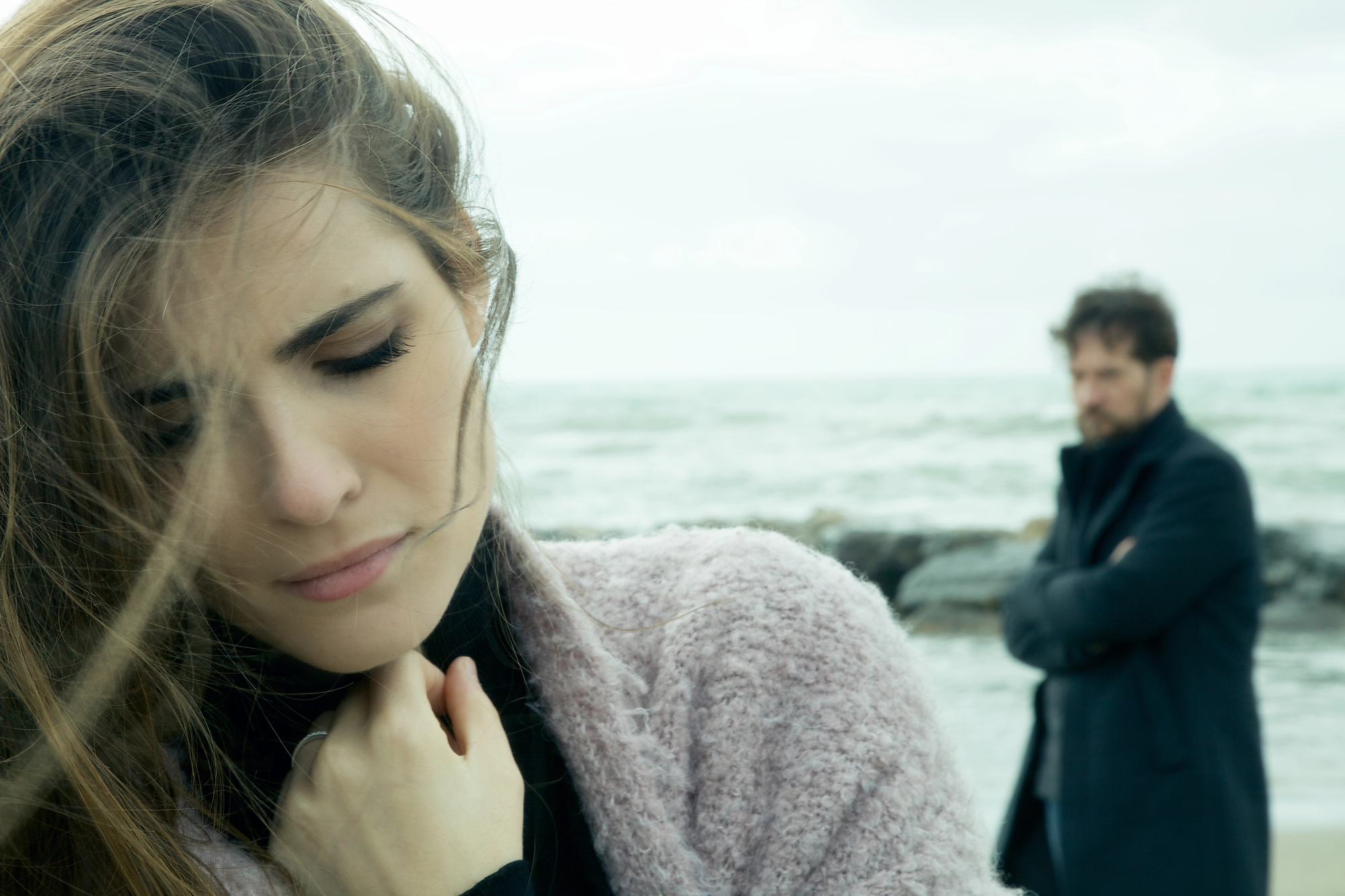 A pensive woman with long hair stands in the foreground near the shore, her eyes closed and hand near her chest. A man with a beard, wearing a dark coat, stands in the background with crossed arms, looking out at the sea. The mood is somber and reflective.