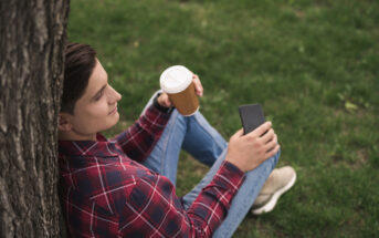 A person wearing a red plaid shirt and blue jeans sits on grass, leaning against a tree. They are holding a to-go coffee cup in one hand and a smartphone in the other, looking content and relaxed. The background is grassy, indicating a park or outdoor space.