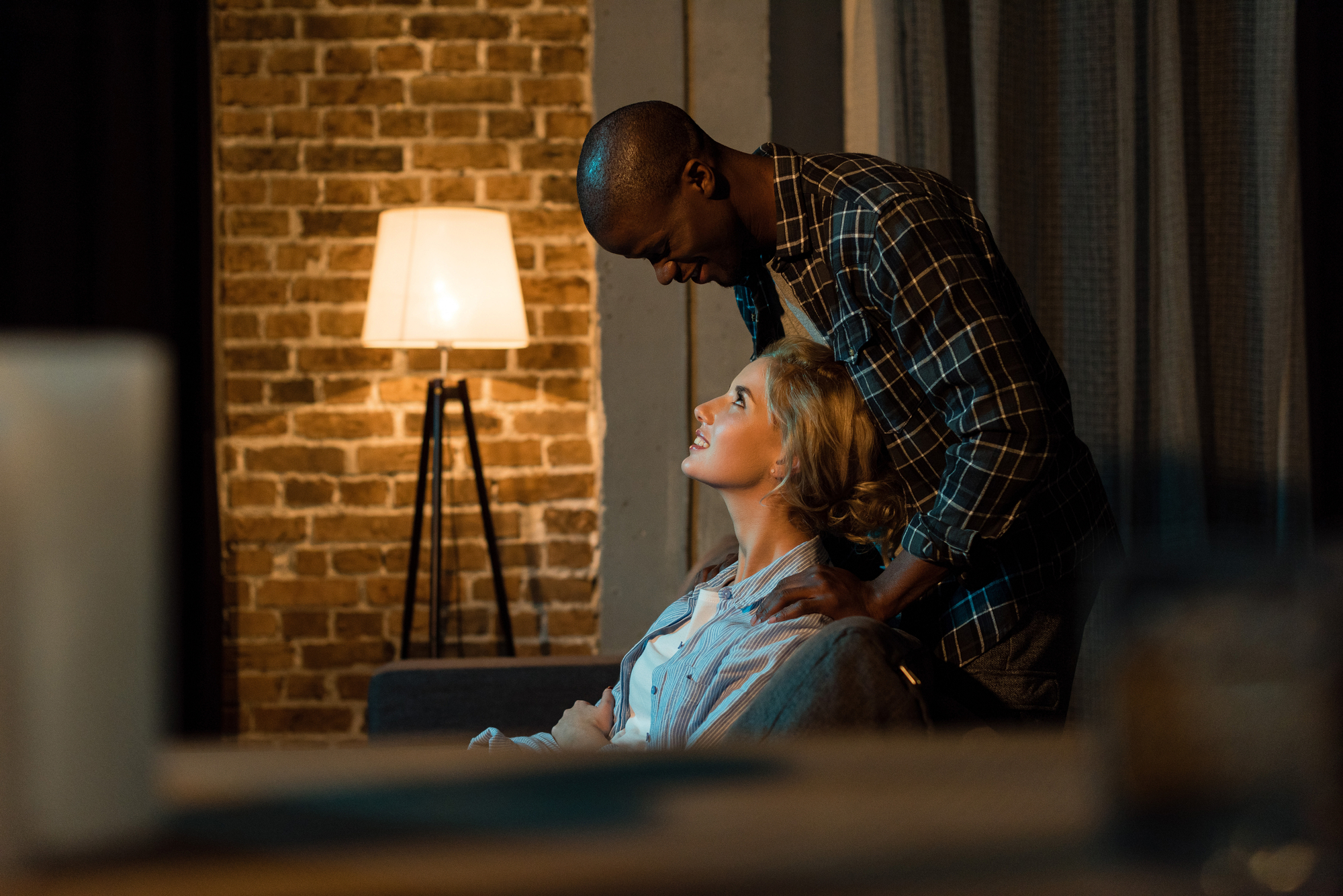 A man and woman share a tender moment in a cozy, dimly lit room with brick walls and a standing lamp. The man stands behind the woman, who is seated, and they gaze into each other's eyes, both smiling warmly.