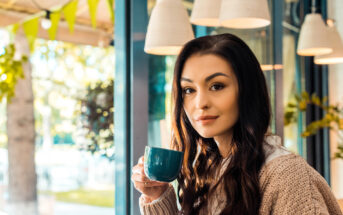 A woman with long dark hair is sitting in a cozy café, holding a teal coffee mug near her face. She is wearing a beige sweater and looking towards the camera. There are hanging pendant lights and green foliage visible in the background through large windows.