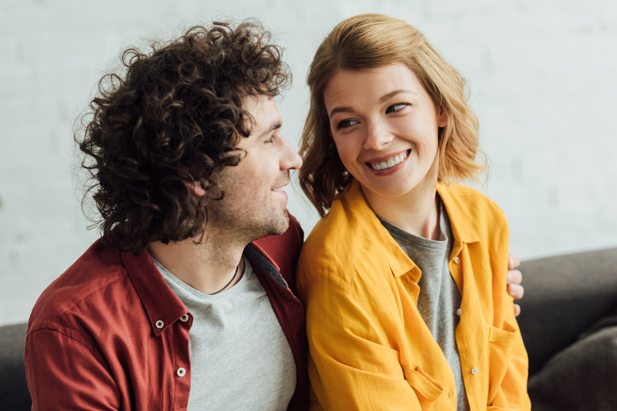 A man with curly hair and a reddish shirt and a woman with shoulder-length blonde hair and a mustard yellow shirt smile warmly at each other while sitting on a couch. They appear happy and engaged in a light-hearted conversation.