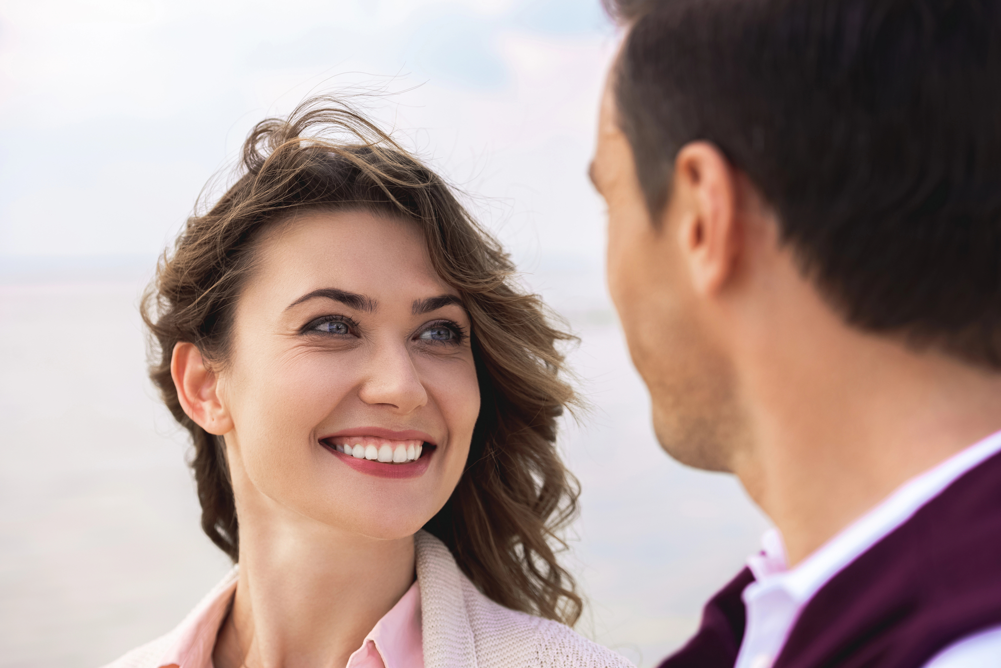 A woman with wavy hair and a bright smile looks at a man who is facing her, with the ocean blurred in the background. The woman wears a light sweater and a pink shirt, and the man wears a dark jacket with a white shirt.