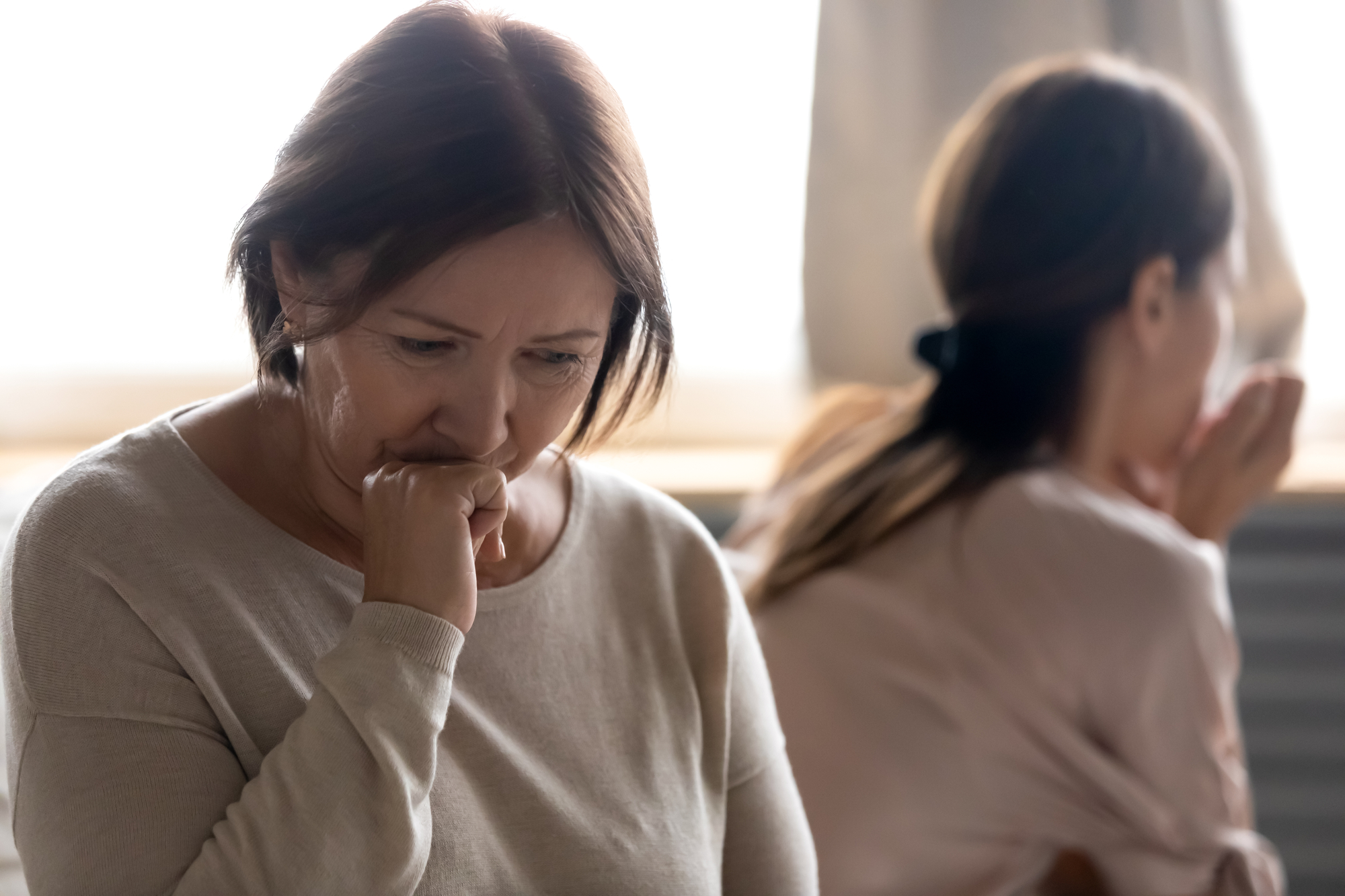 Two women are depicted in the image. In the foreground, an older woman with short brown hair looks down thoughtfully, with her fist against her mouth. In the background, a younger woman, facing away, appears to be deep in thought as well.