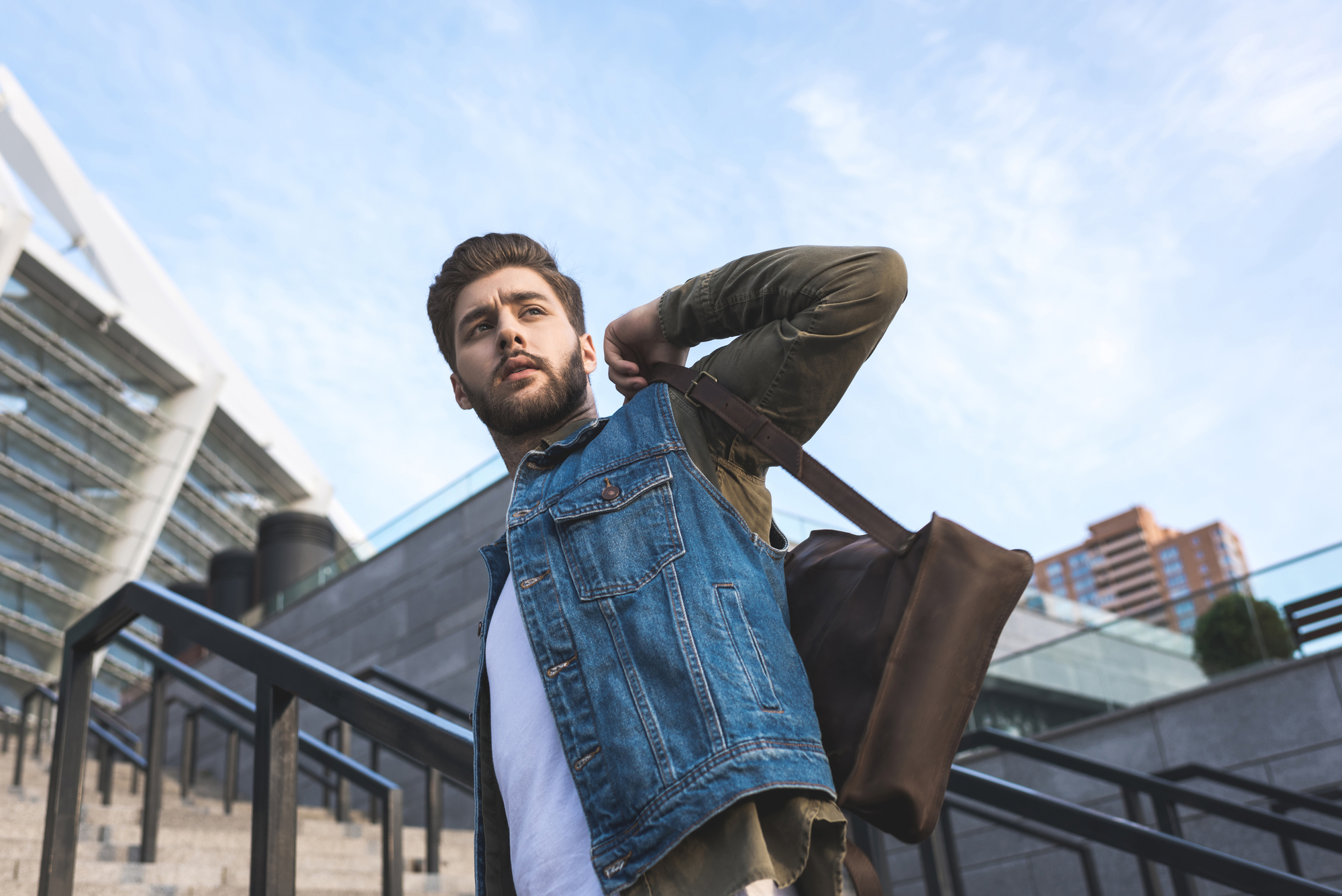 A man with a beard, wearing a blue denim jacket over a green shirt, carries a large brown bag over his shoulder as he stands on an outdoor staircase. Modern buildings and the sky with scattered clouds are visible in the background.