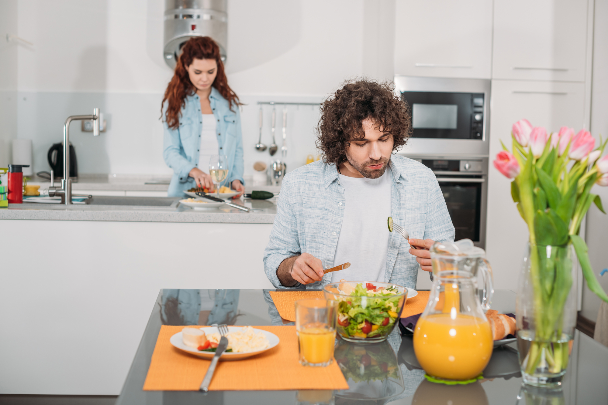 A man with curly hair sits at a dining table eating a salad, while a woman in the background stands by the kitchen sink holding a plate. The table also has a pitcher of orange juice, a glass of juice, and a vase with pink tulips.