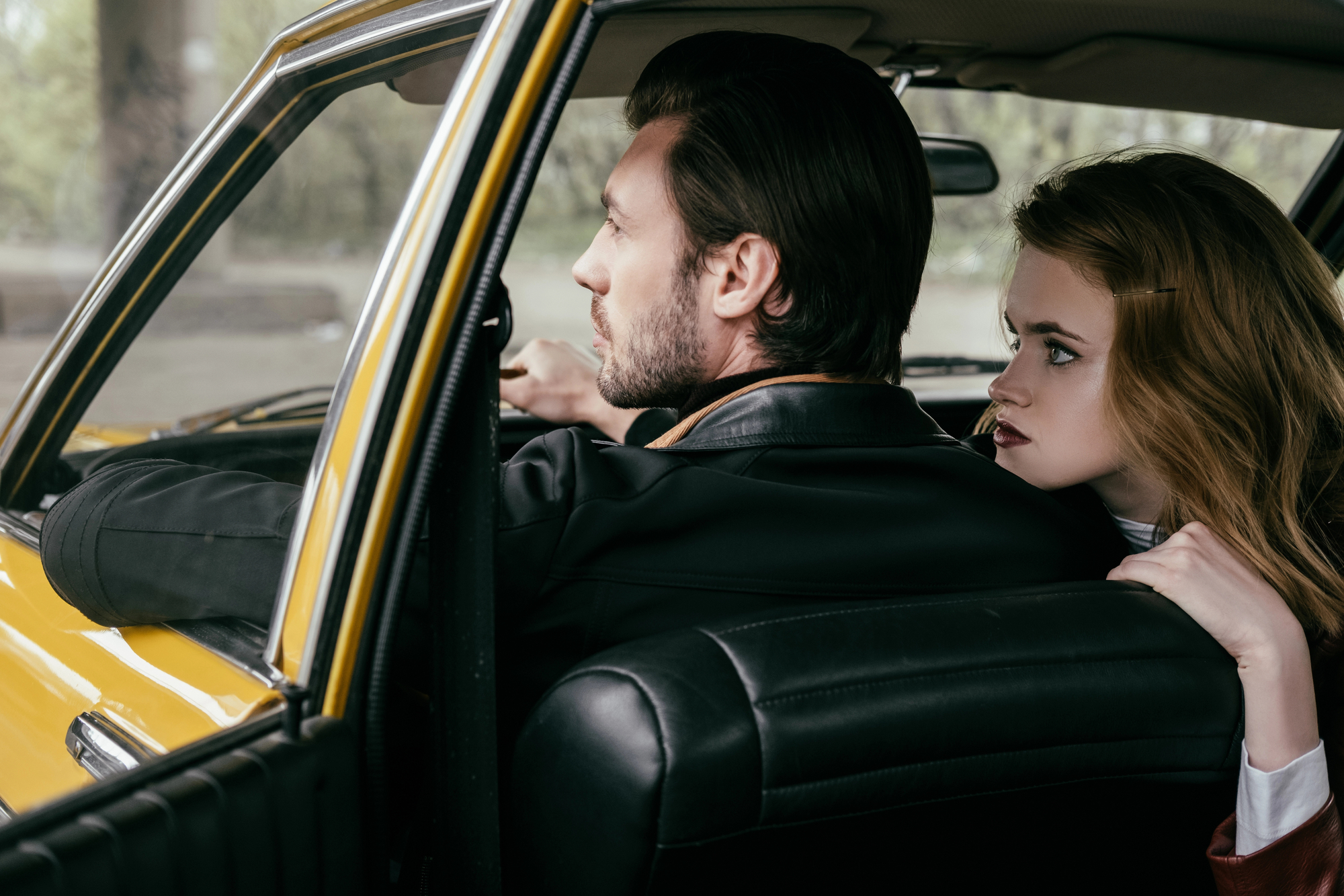 A man is driving a yellow car, looking focused and wearing a black leather jacket. A woman with long hair, seated in the back, leans forward and looks at him with a concerned expression. The interior of the car is black, with a blurred outdoor background.