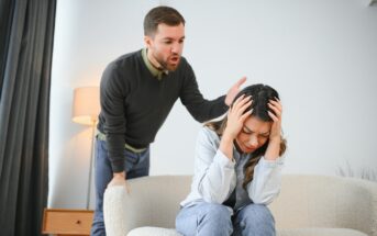 A man with a beard is shouting at a woman sitting on a sofa. The woman, distressed, is holding her head in her hands. The setting is a modern living room with a lamp and side table in the background.