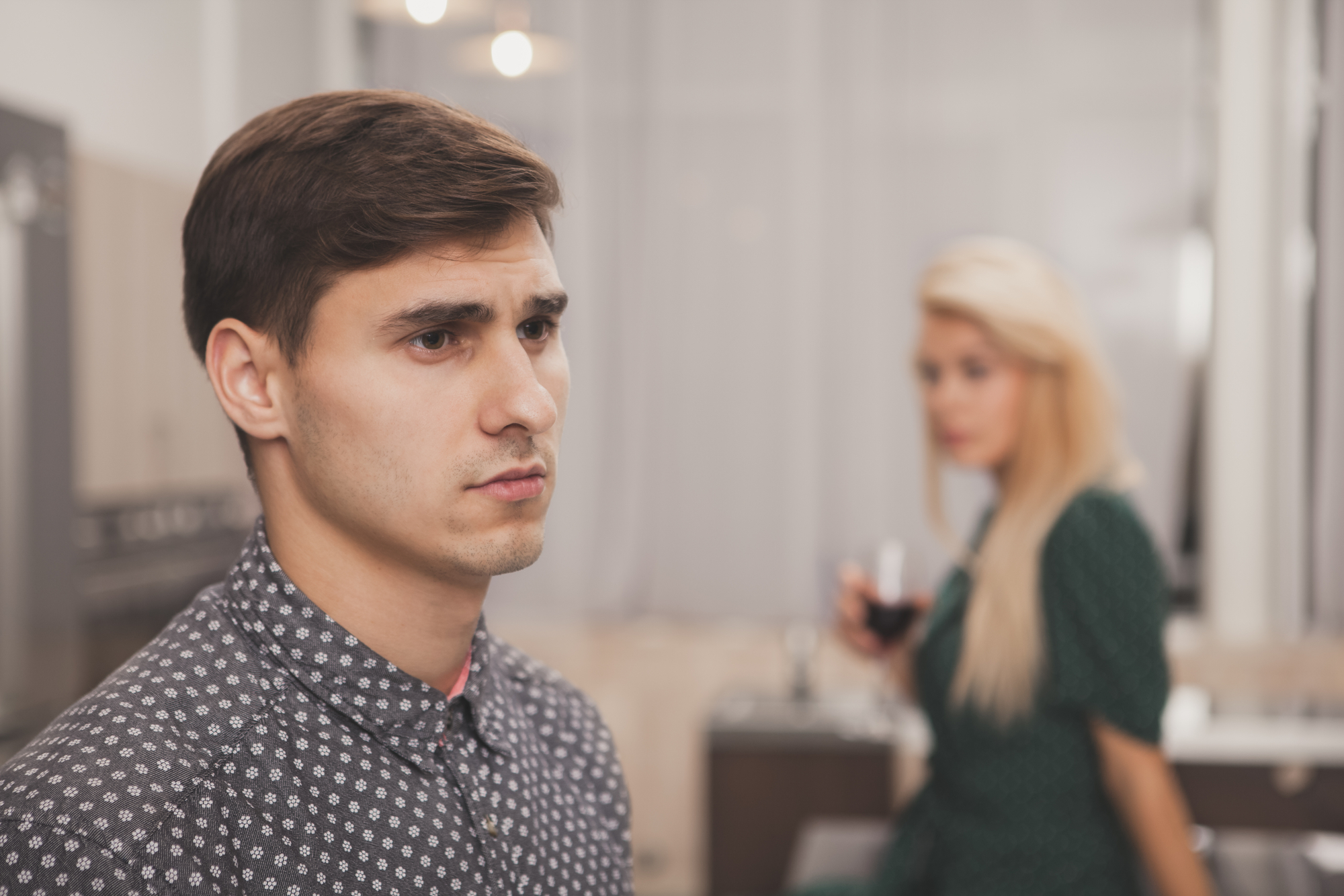 A young man with short brown hair looks serious and introspective, standing indoors in a well-lit room. In the background, a blonde woman in a green dress holds a glass of wine and looks in his direction, though distant and slightly out of focus.