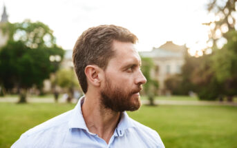 A man with short, curly hair and a beard, wearing a light blue shirt, gazes to his left. He is standing outdoors in a lush green park with trees and buildings in the background, under a partly cloudy sky.