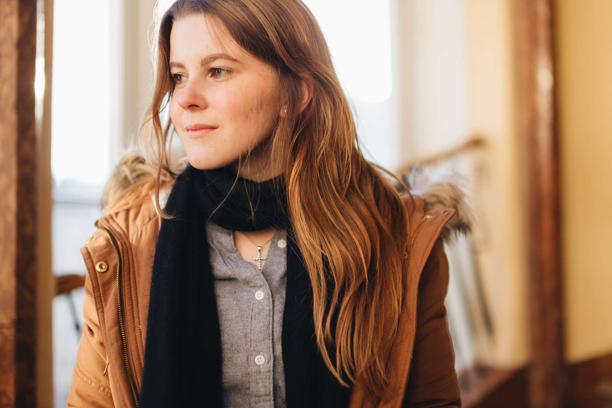 A young woman with long, light brown hair is looking to the side. She is wearing a brown jacket with a fur-trimmed hood, a black scarf, and a grey shirt. The background features blurred indoor elements with warm lighting.