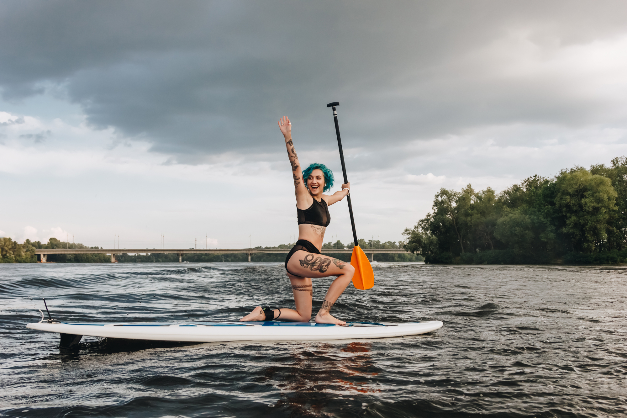 Person with blue hair is kneeling on a paddleboard in a body of water, wearing a black bikini and holding a paddle. They are smiling with one hand raised. The sky is mostly cloudy, and the background features a bridge and trees along the shoreline.