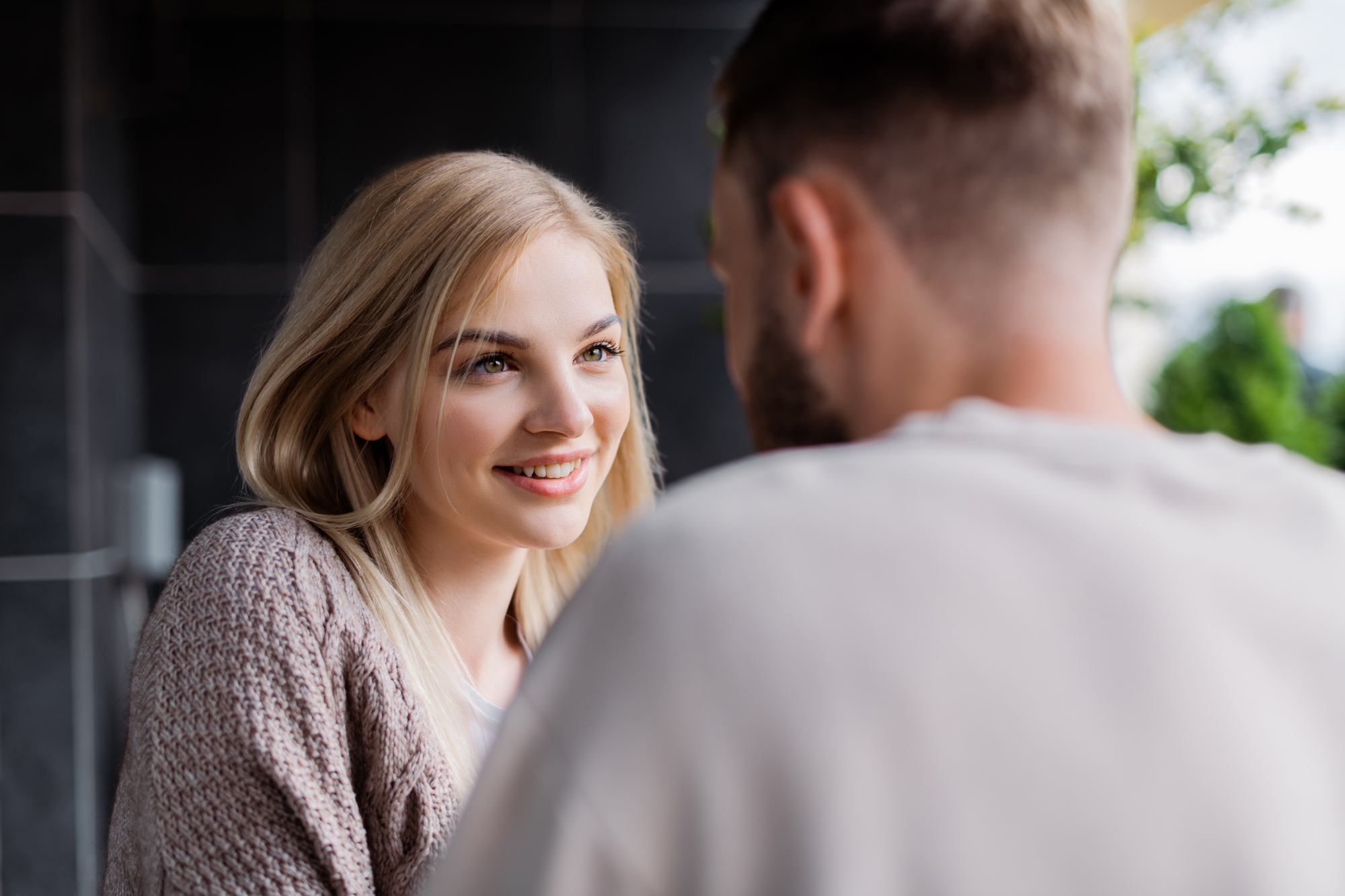 A woman with long blonde hair smiles warmly at a man with short hair in an outdoor setting. She is wearing a knit sweater and looking at him intently. The background is slightly blurred, with some greenery visible.