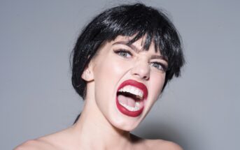 A person with short black hair and bold red lipstick is expressing anger or aggression. Their mouth is open wide, showing teeth, and their eyes are wide open, conveying an intense emotion. The background is plain and neutral.