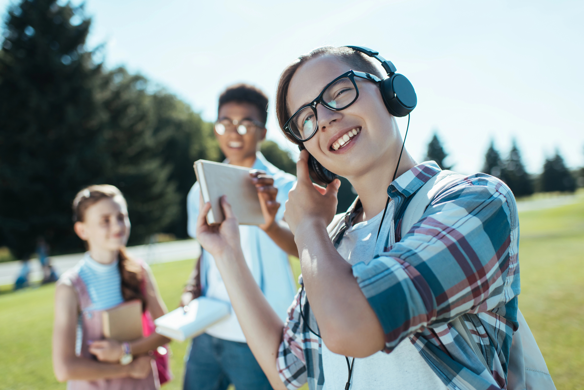 A young person with glasses and headphones smiles while holding a tablet and touching their ear. Two friends stand in the background, one holding a book and the other looking at a tablet. They are outside on a sunny day with trees in the background.