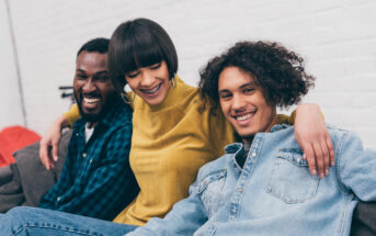 Three friends are sitting on a couch, smiling and laughing. The person on the left wears a plaid shirt, the person in the middle, with short hair and hoop earrings, wears a yellow sweater, and the person on the right, with curly hair, wears a denim shirt.