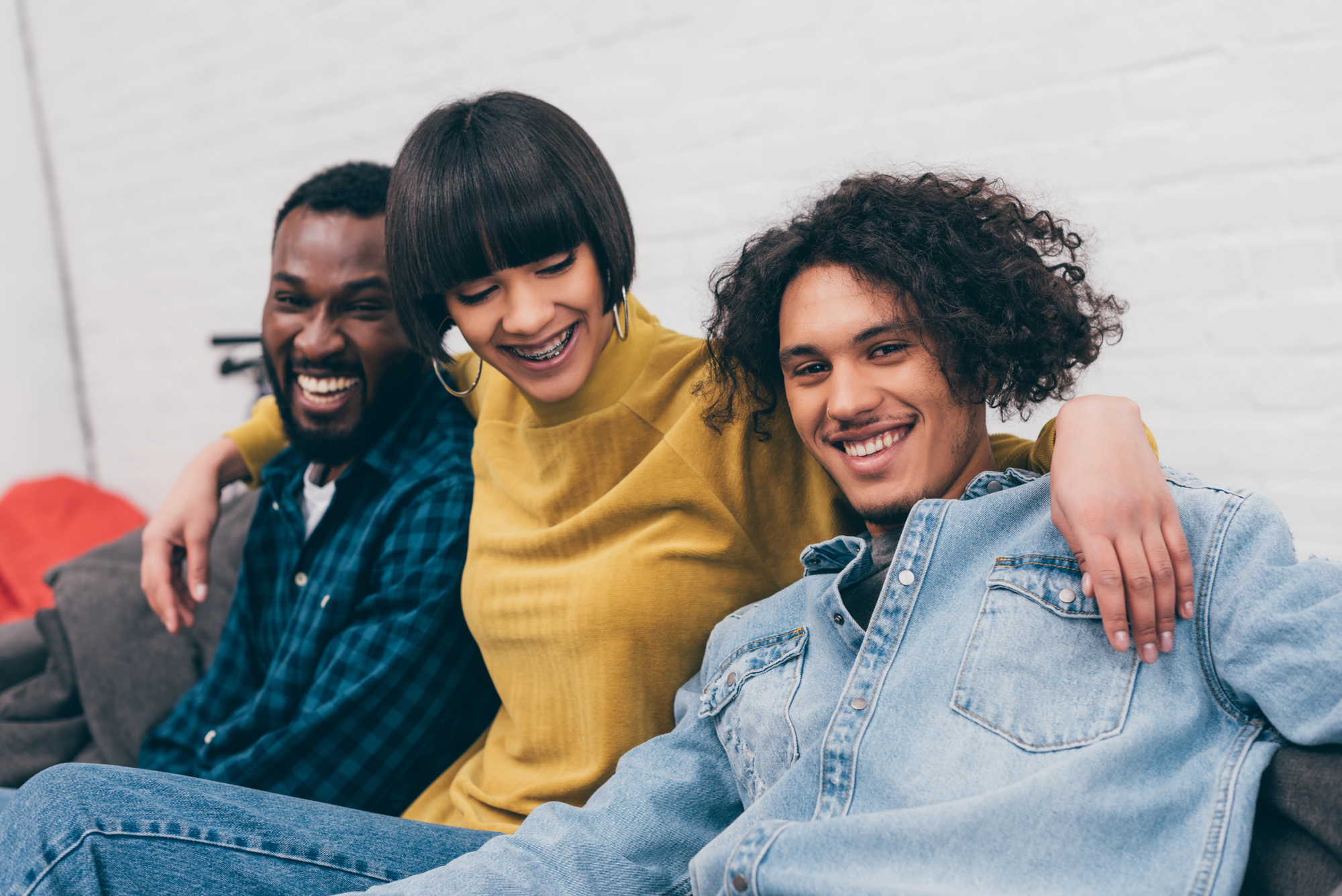 Three friends are sitting on a couch, smiling and laughing. The person on the left wears a plaid shirt, the person in the middle, with short hair and hoop earrings, wears a yellow sweater, and the person on the right, with curly hair, wears a denim shirt.