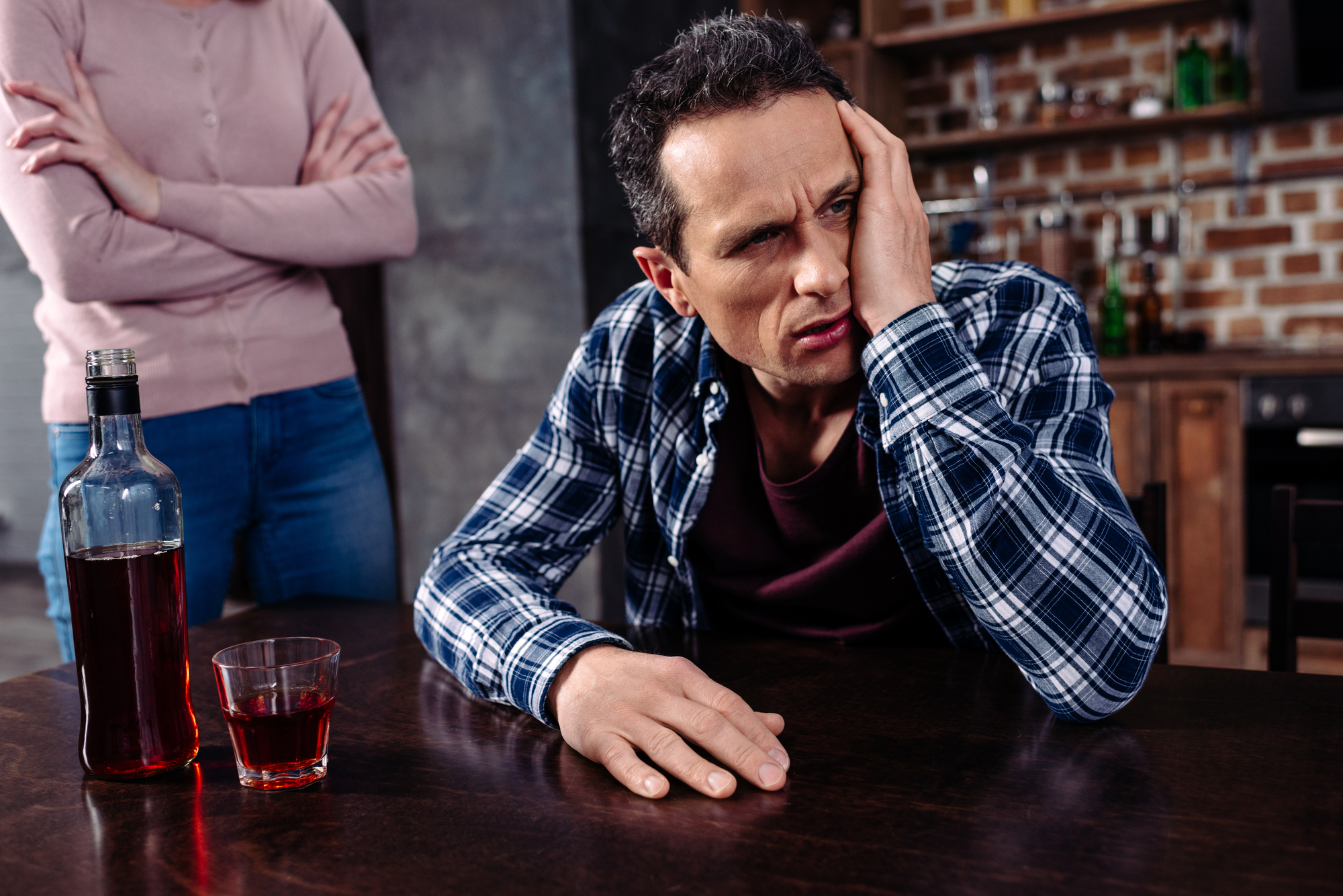 A man sits at a table with a distressed expression, resting his head on one hand. A bottle and a glass of liquor are in front of him. In the background, a person stands with arms crossed, partially visible from the chest down. The scene appears tense.