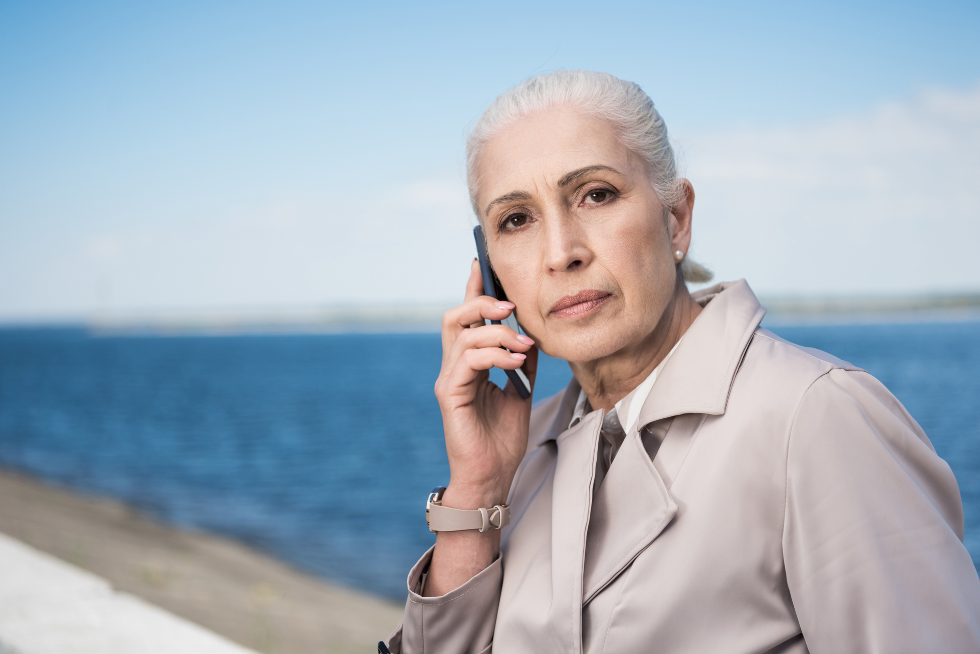 A woman with grey hair in a light trench coat is holding a phone to her ear. She has a serious expression and is standing outdoors near a body of water with a blue sky in the background.