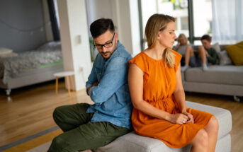 A woman in an orange dress and a man in a denim shirt sit back to back on a couch, looking upset. Two children sit on another couch in the background, focusing on a tablet. The room has wooden floors and natural light from large windows.