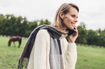 A smiling woman with a gray scarf and cream sweater is talking on her phone in a grassy field. A horse grazes in the background, with trees lining the edge of the field.