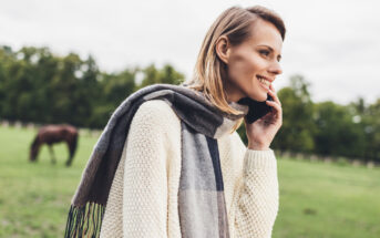 A smiling woman with a gray scarf and cream sweater is talking on her phone in a grassy field. A horse grazes in the background, with trees lining the edge of the field.