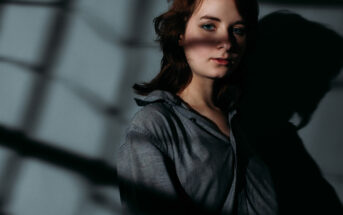 A person with shoulder-length hair stands against a wall with shadows cast over them. They are wearing a loose, button-up shirt and looking pensively at the camera. The lighting creates a dramatic contrast with the background.