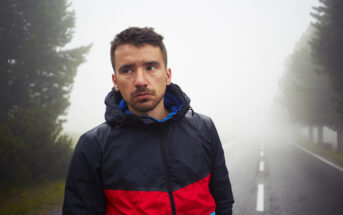 A man in a red and black jacket stands on an empty, foggy road surrounded by tall trees. He looks pensively into the distance. The atmosphere is misty, and visibility is low.