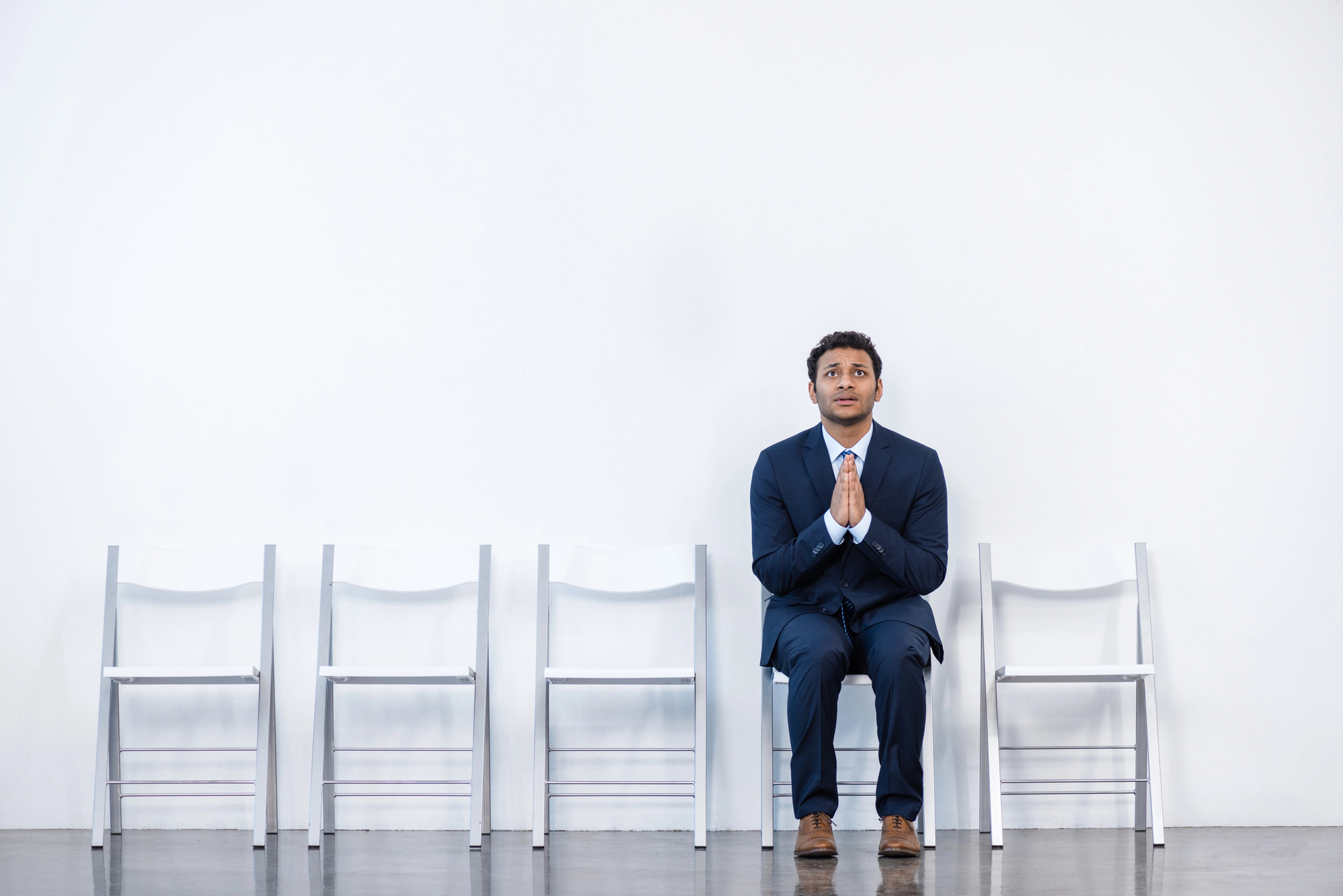 A man in a blue suit sits with his hands clasped together on one of five white chairs against a white wall. He looks slightly upward, appearing anxious or hopeful. The other four chairs are empty, and the floor is a shiny, polished surface.