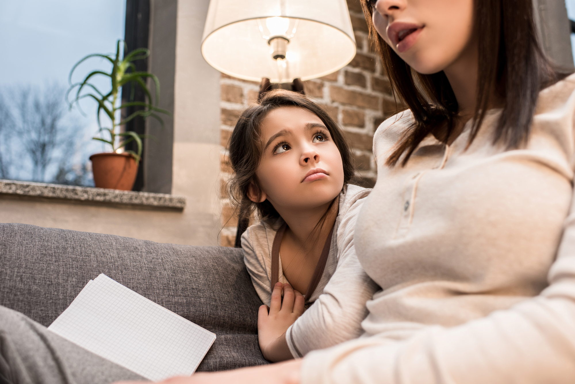 A young girl with long hair looks up at a woman sitting beside her on a sofa, holding a piece of grid paper. The woman appears to be speaking, and both are in a cozy, warmly lit room with a potted plant and a lamp in the background.