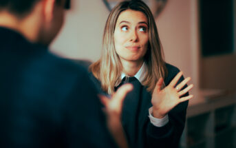 A woman with shoulder-length blonde hair is sitting indoors, raising her hands with an expressive look while talking to a person facing her. She wears a dark grey sweater over a collared shirt. The room has a modern decor.