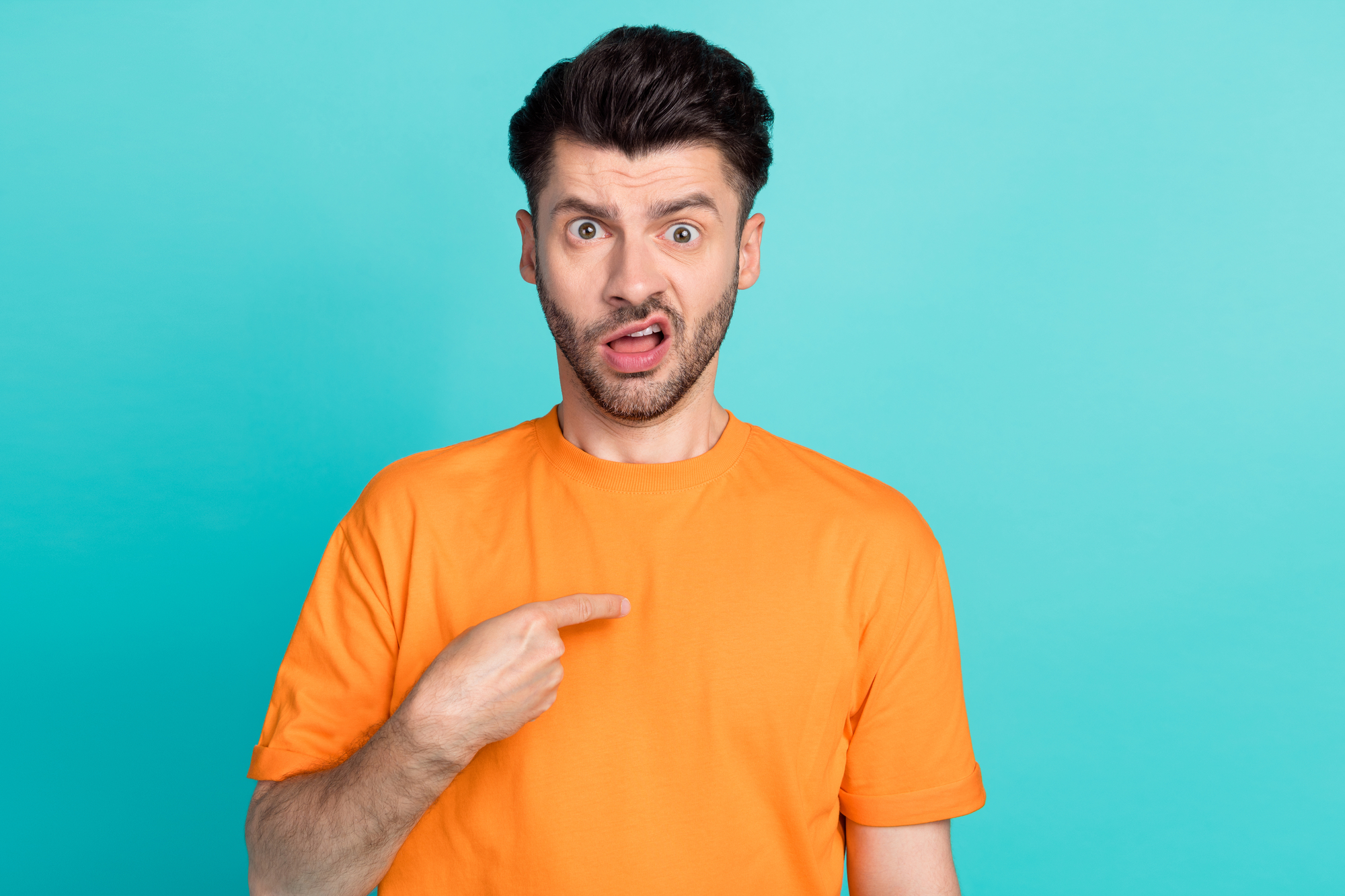 A man with short dark hair and a beard is standing against a blue background, wearing an orange t-shirt. He has a surprised expression on his face, with his eyebrows raised and his mouth slightly open. He is pointing at himself with his right hand.