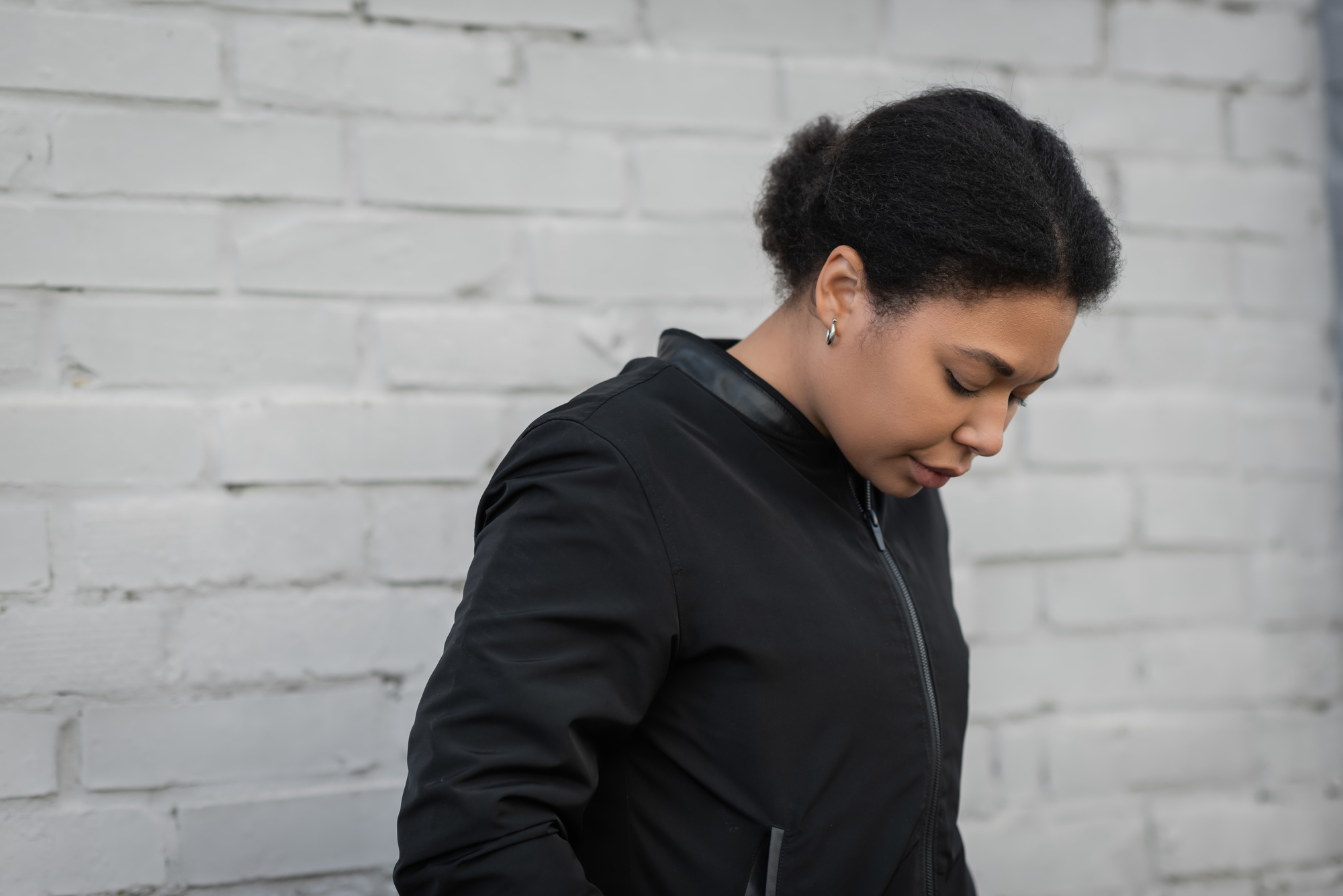 A person with short, curly hair wearing a black jacket looks down while standing in front of a light gray brick wall.