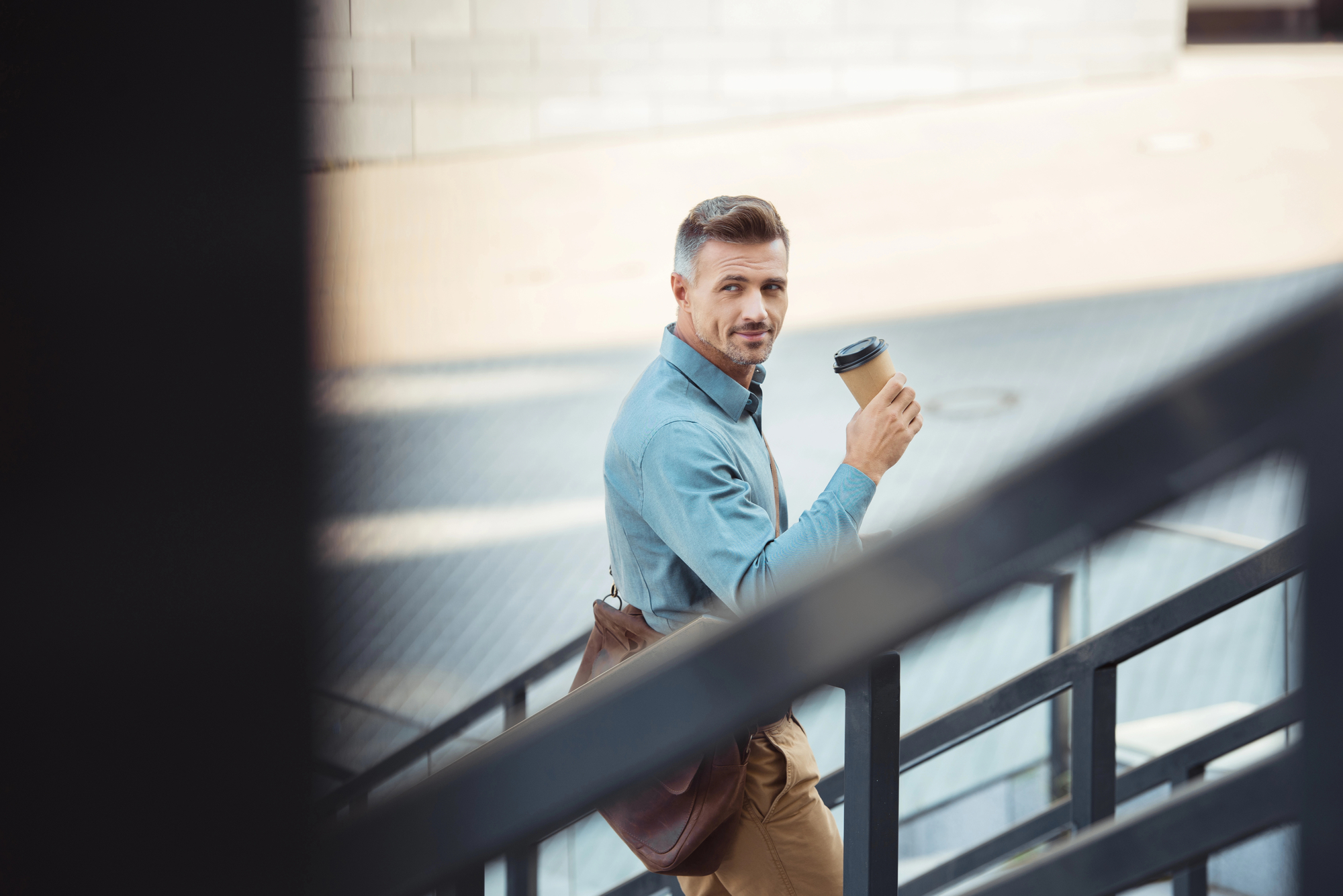 A man in a blue shirt holding a coffee cup ascends a set of stairs. He has a brown shoulder bag and is looking over his shoulder, smiling slightly. The background is urban, featuring a light-colored building and paved ground.