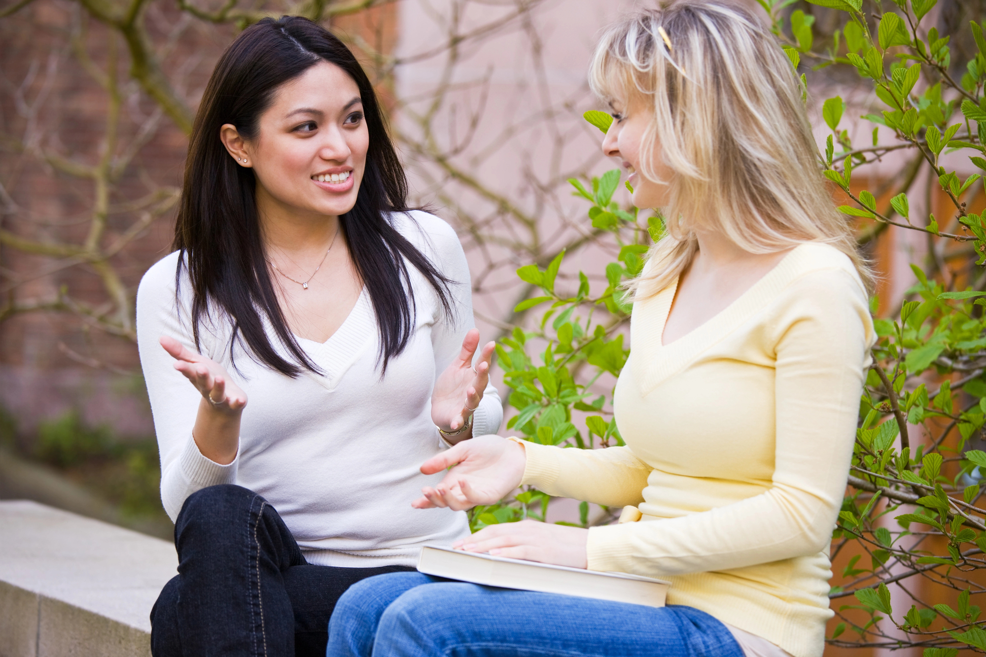 Two women are sitting and engaged in a lively conversation outdoors. One has long black hair and is wearing a white sweater, while the other has blonde hair and is dressed in a yellow sweater, holding a book on her lap. Green foliage and building structures are in the background.