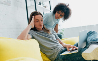 A person with long hair sits on a yellow couch using a laptop, looking concerned. Another person with curly hair stands next to them, leaning over to see the laptop screen, appearing focused and inquisitive. A white, bright room is in the background.