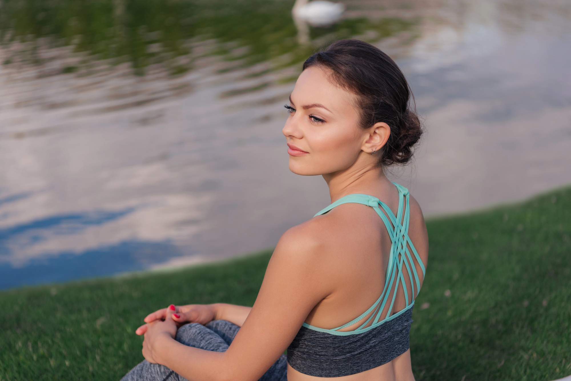 A young woman with dark hair tied back is sitting on the grass near a body of water. She is wearing a teal sports bra and gray leggings. Her gaze is directed slightly to the side, and she appears calm and relaxed. The background includes a swan in the water.