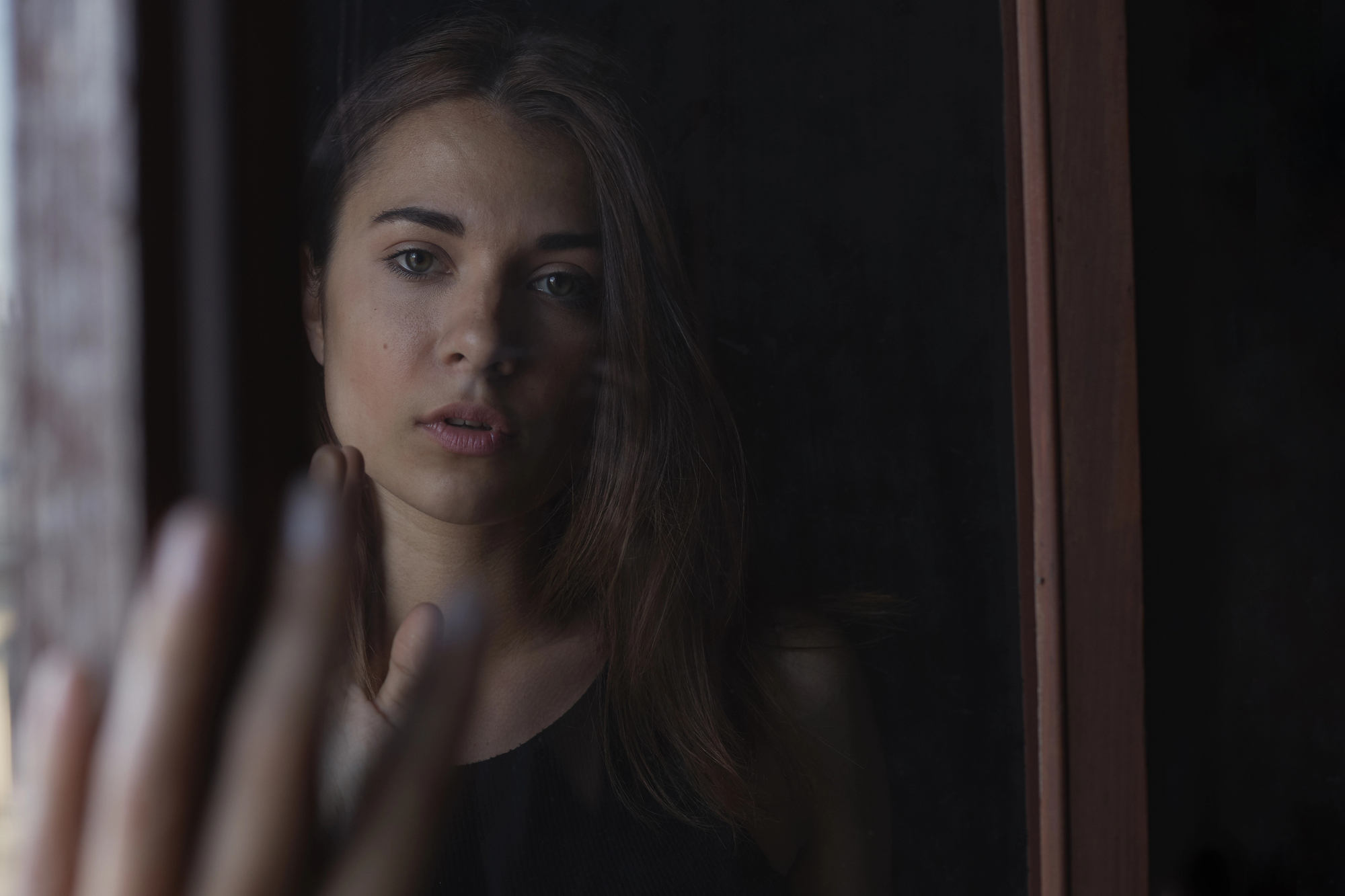 A woman with long brown hair and a neutral expression gazes through a window. Her hand is raised and pressed against the glass. She wears a dark, sleeveless top and is surrounded by dim lighting. The scene conveys a sense of contemplation.