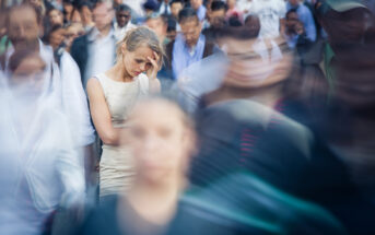 A woman stands still in a crowded area with a distressed expression, hand on her forehead. The surrounding crowd appears in motion blur, emphasizing her stationary and troubled state amidst the rush of people. The image conveys a sense of isolation and overwhelm.