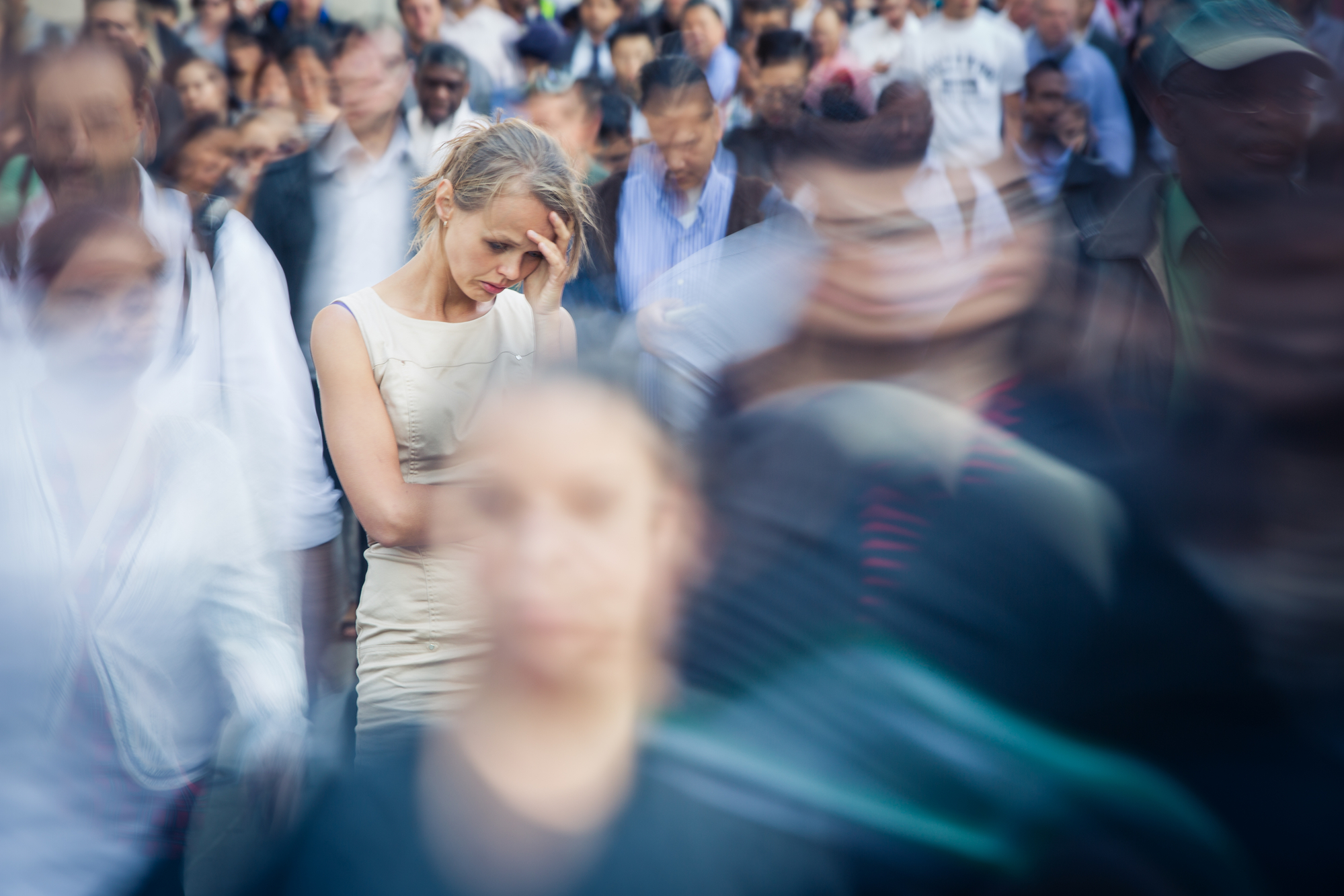 A woman stands still in a crowded area with a distressed expression, hand on her forehead. The surrounding crowd appears in motion blur, emphasizing her stationary and troubled state amidst the rush of people. The image conveys a sense of isolation and overwhelm.