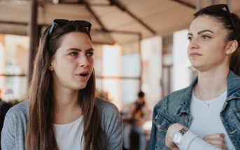 Two women are seated and engaged in conversation at an outdoor café. The woman on the left is speaking, while the woman on the right listens attentively with a thoughtful expression. Both have long hair and are casually dressed, with sunglasses resting on their heads.