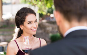 A woman with dark hair smiles while holding a glass of red wine, looking at a man with his back to the camera. The scene is outdoors with greenery in the background. The woman is wearing a sleeveless top and large hoop earrings.