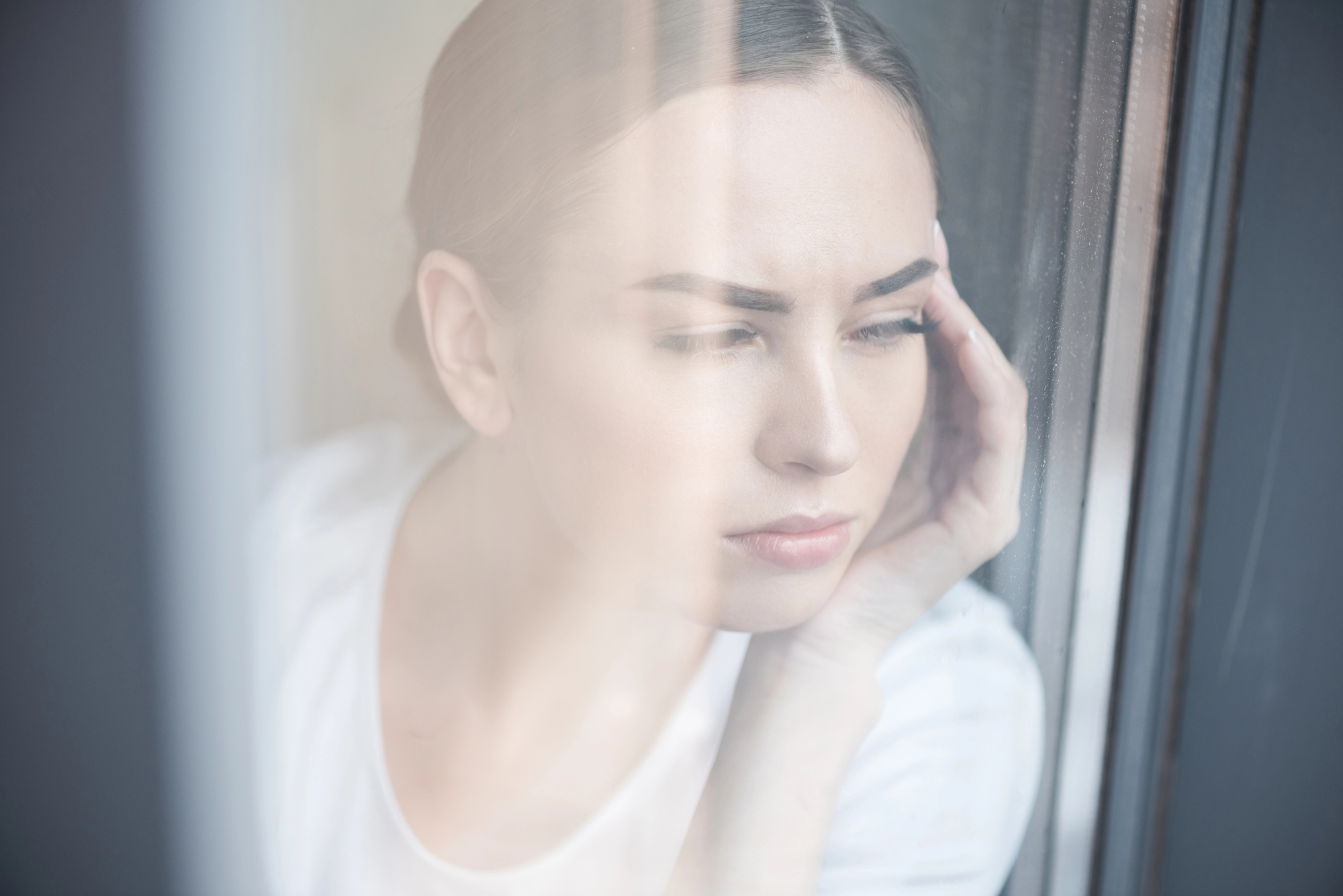 A woman with dark hair tied back is gazing out of a window, resting her face on her hand. She appears thoughtful and pensive. The photo is taken through the glass, adding a soft, reflective quality to the image.