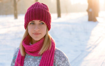 A young woman stands outdoors on a snowy day, wearing a pink knitted hat and matching scarf. She has blonde hair and is dressed in a geometrically patterned sweater. Sunlight filters through trees in the background, casting a warm glow.